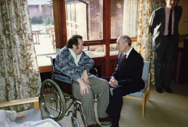 Leonard Cheshire talking to a man in a wheelchair by a window inside a lounge