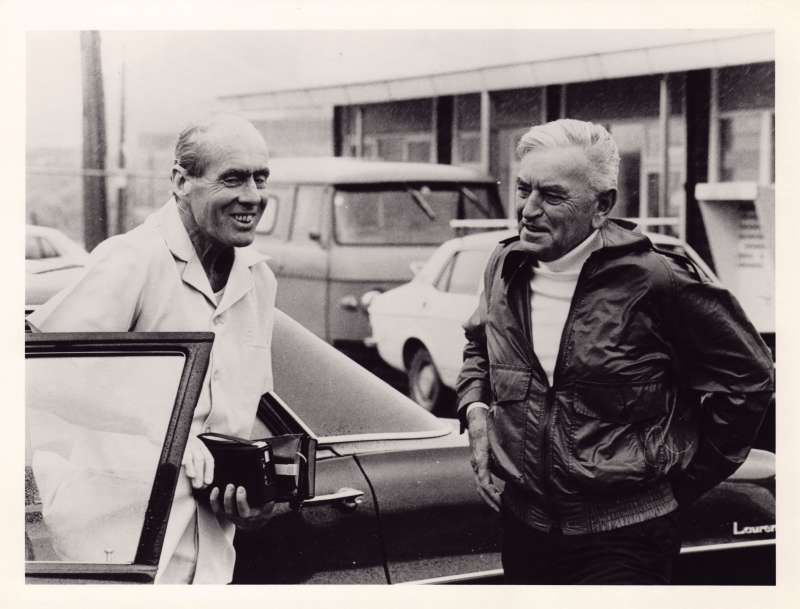 Leonard Cheshire standing near a car talking with another man in a leather jacket on a windy day