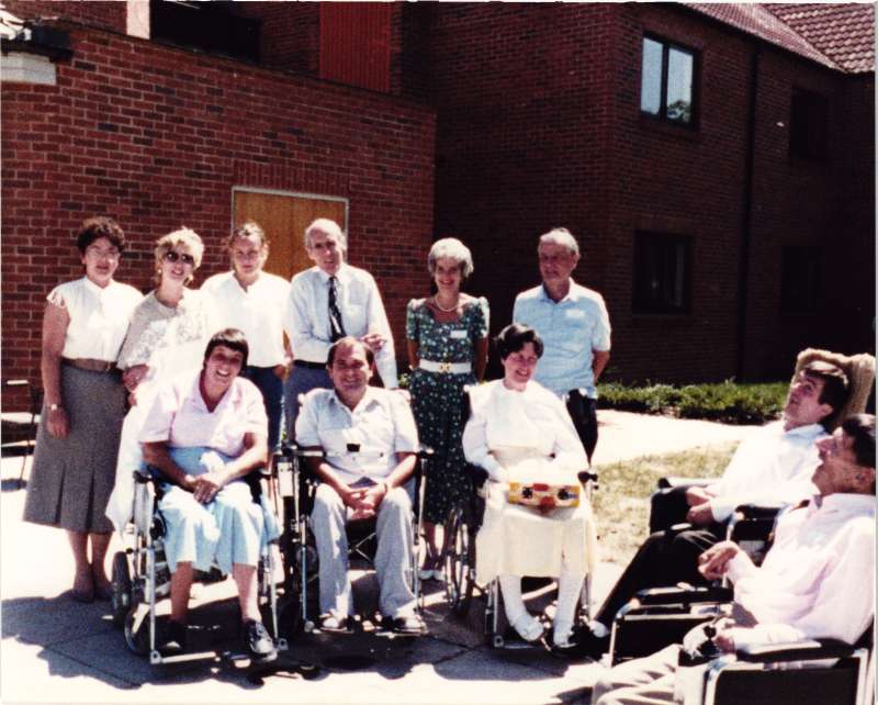 An older Leonard Cheshire with a group of people, some in wheelchairs in front of a brick building