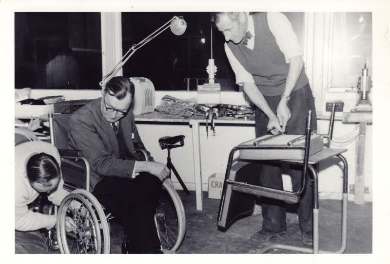 A man sat in a wheelchair whilst another man adjusts the wheel mechanism. Another man altering a wheelchair seat