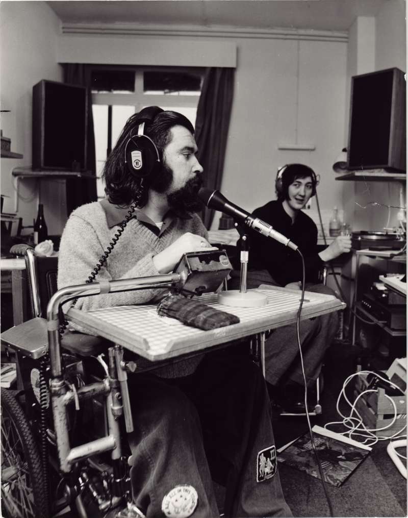 A man in a wheelchair with headphones and microphone in a home-based recording studio, with another person wearing headphones in the background