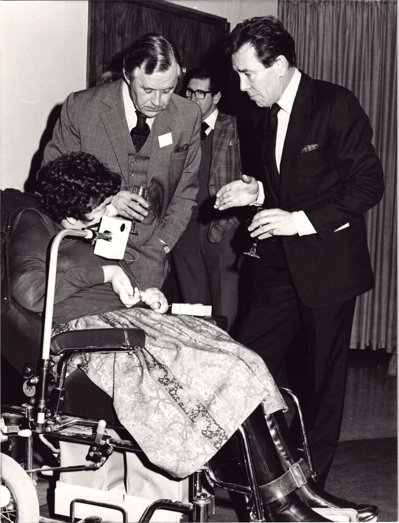 A woman in a wheelchair talking with two men in suits holding wine glasses