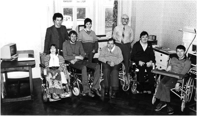 Five people in wheelchairs and three others standing in a room with computers in the background
