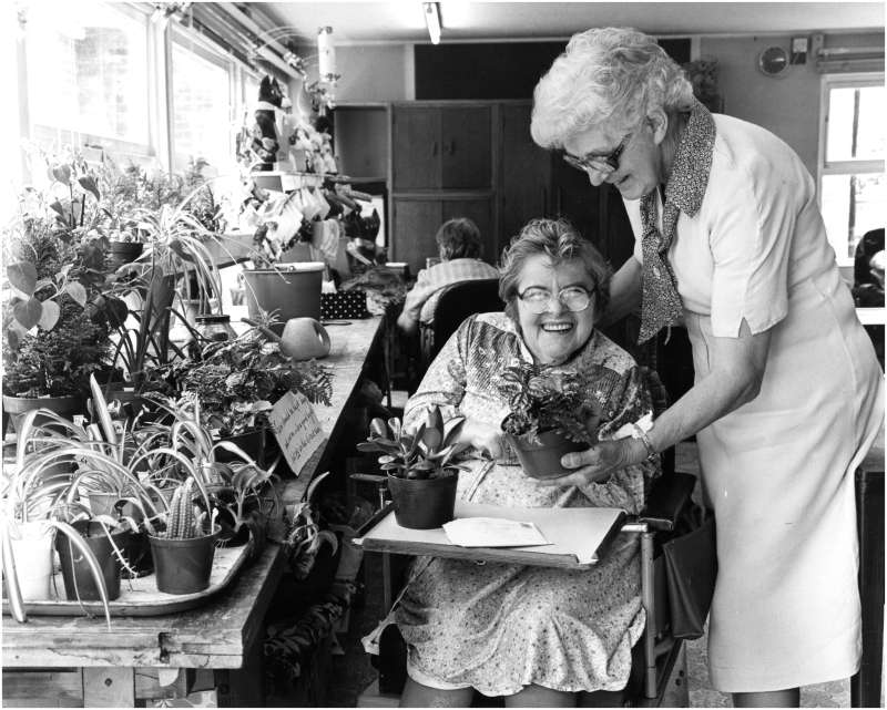 A smiling lady in a wheelchair holding two pot plants, sat next to a plant bench, with a lady helping her