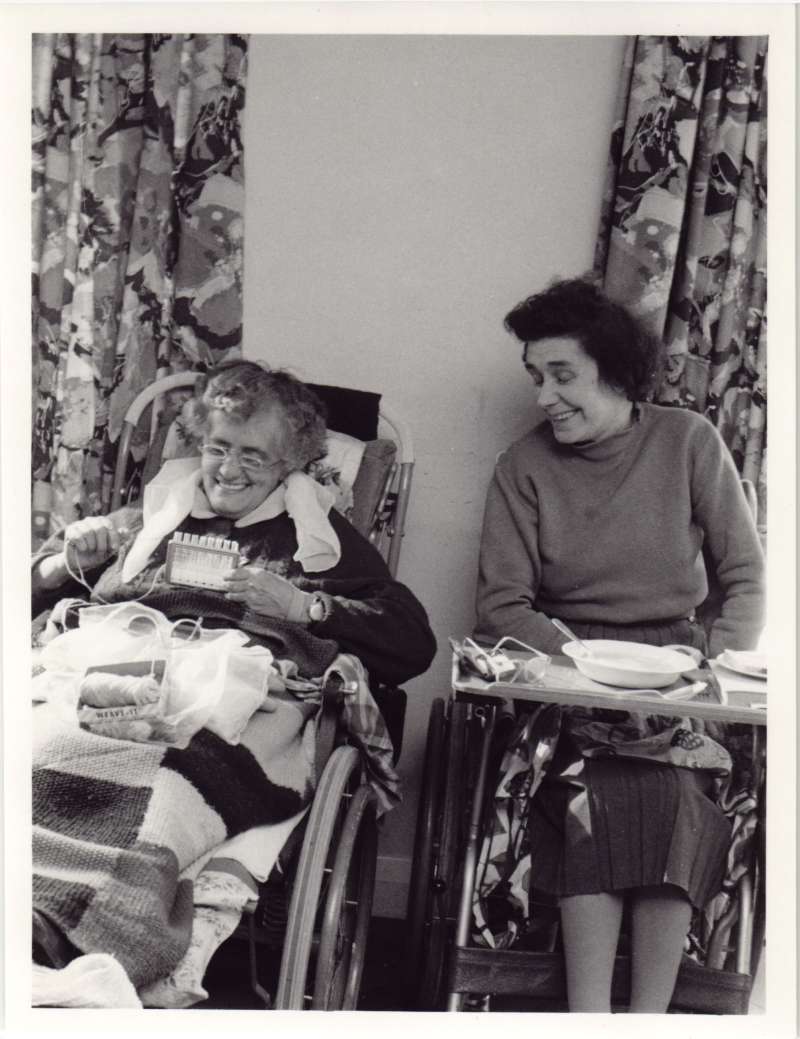 Two women in wheelchairs laughing. One doing needlework, one with a food bowl on a table across her chair