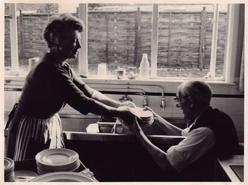 Woman standing, handing a pile of dishes to a man in a wheelchair in front of a kitchen sink