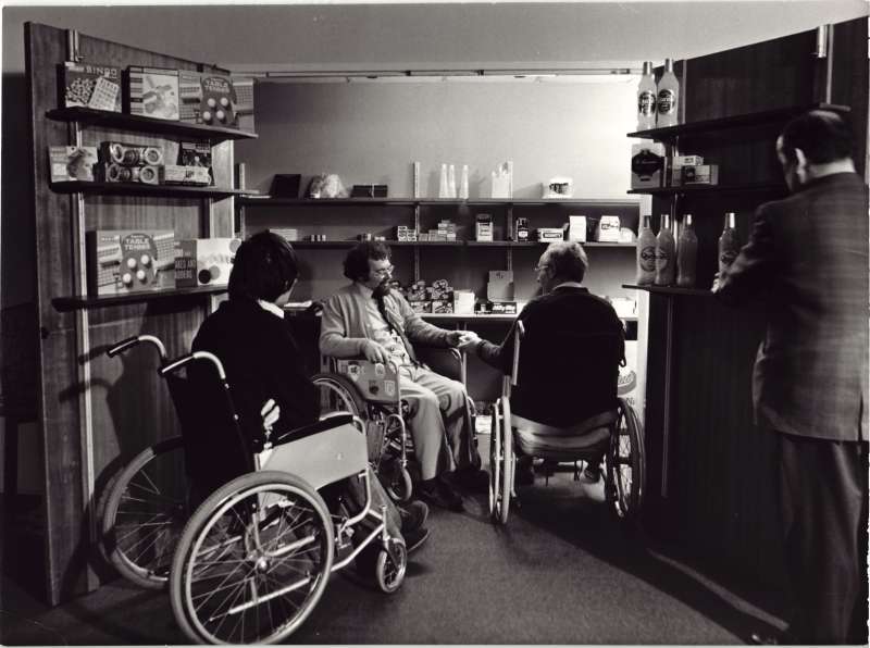 Three men in wheelchairs in a room with shelves stocked with shop supplies