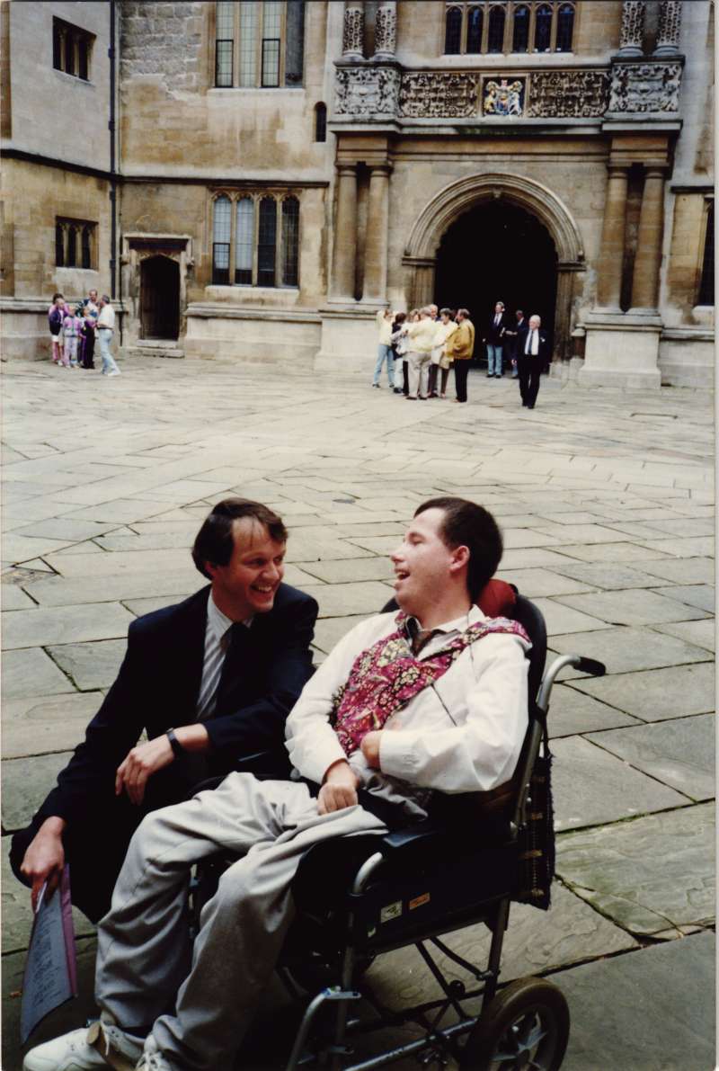 Kevin Whately, actor, crouched down talking to a man in a wheelchair in a courtyard area