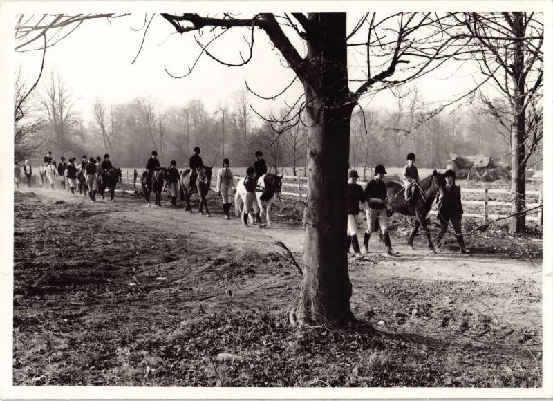 Photo of several people out for a horse ride in the countryside with others leading horses and standing nearby
