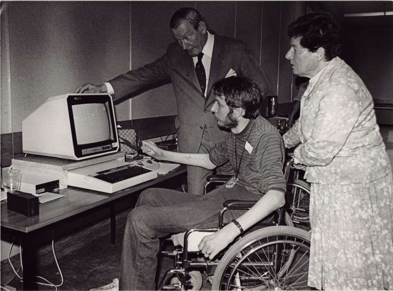 Man in a wheelchair at a computer, with a man and woman watching on