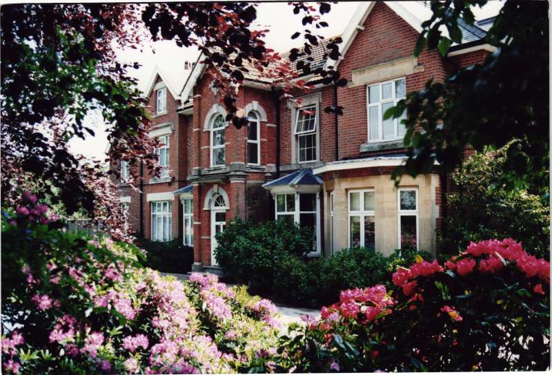 Colour photo of the Heatherley building, taken through trees, with pink and purple flowers in the foreground