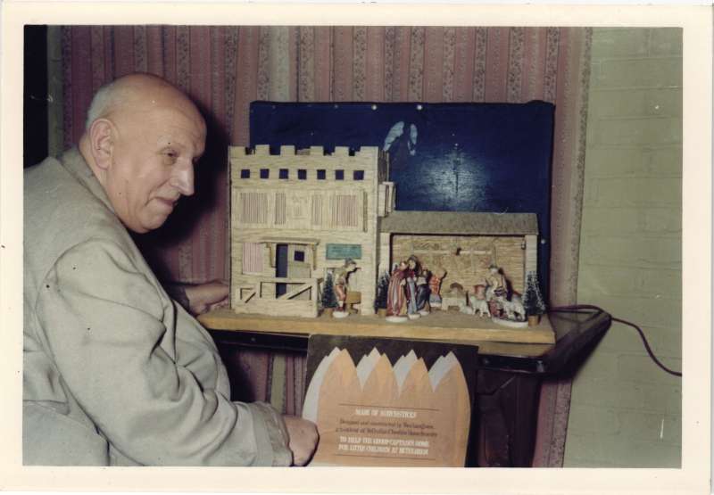 A man in a light grey jacket next to a crib and stable made from from matchsticks