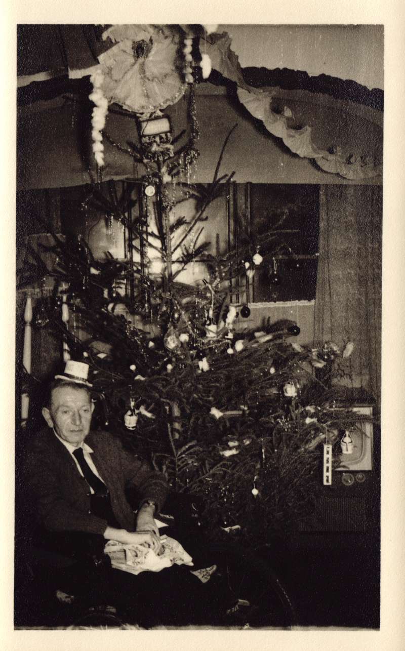 A man in a wheelchair, wearing a mini top hat, sat next to a decorated Christmas tree