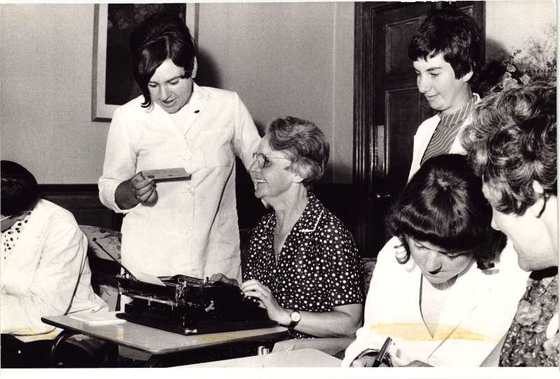 Lady in a black and white spotty top with a typewriter has a letter read to her by another lady