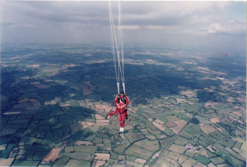 Two men in red parachute jumpsuits and blue hats high above the ground after a parachute jump