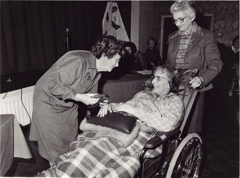 Older lady in a wheelchair with a black handbag on her lap, receiving an award from another lady