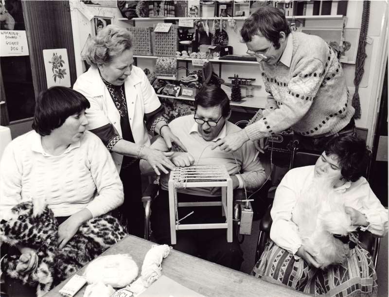 Male resident making a wooden stool with assistance from staff, with two female residents watching on