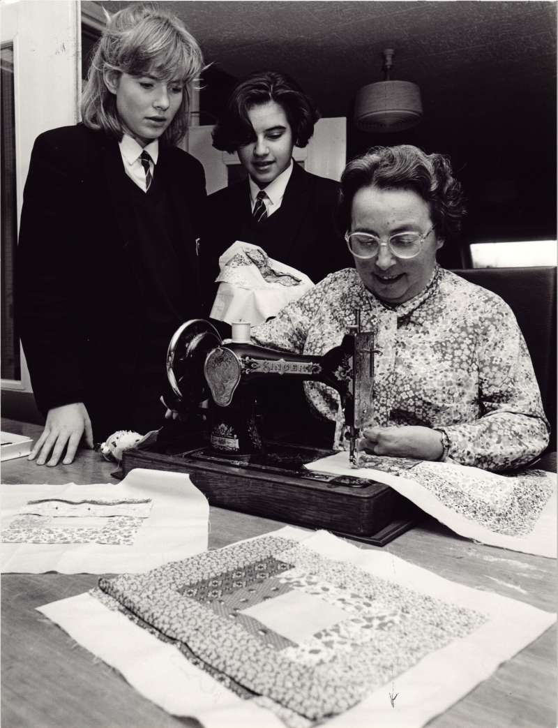 Female resident using a sewing machine, watched on by two schoolgirl volunteers