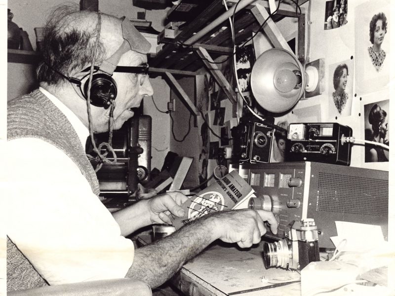 An older man wearing headphones sat at a desk with various amateur radio broadcasting equipment