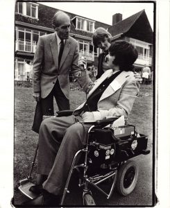 A man in a wheelchair interviewing a man and woman outside a large house