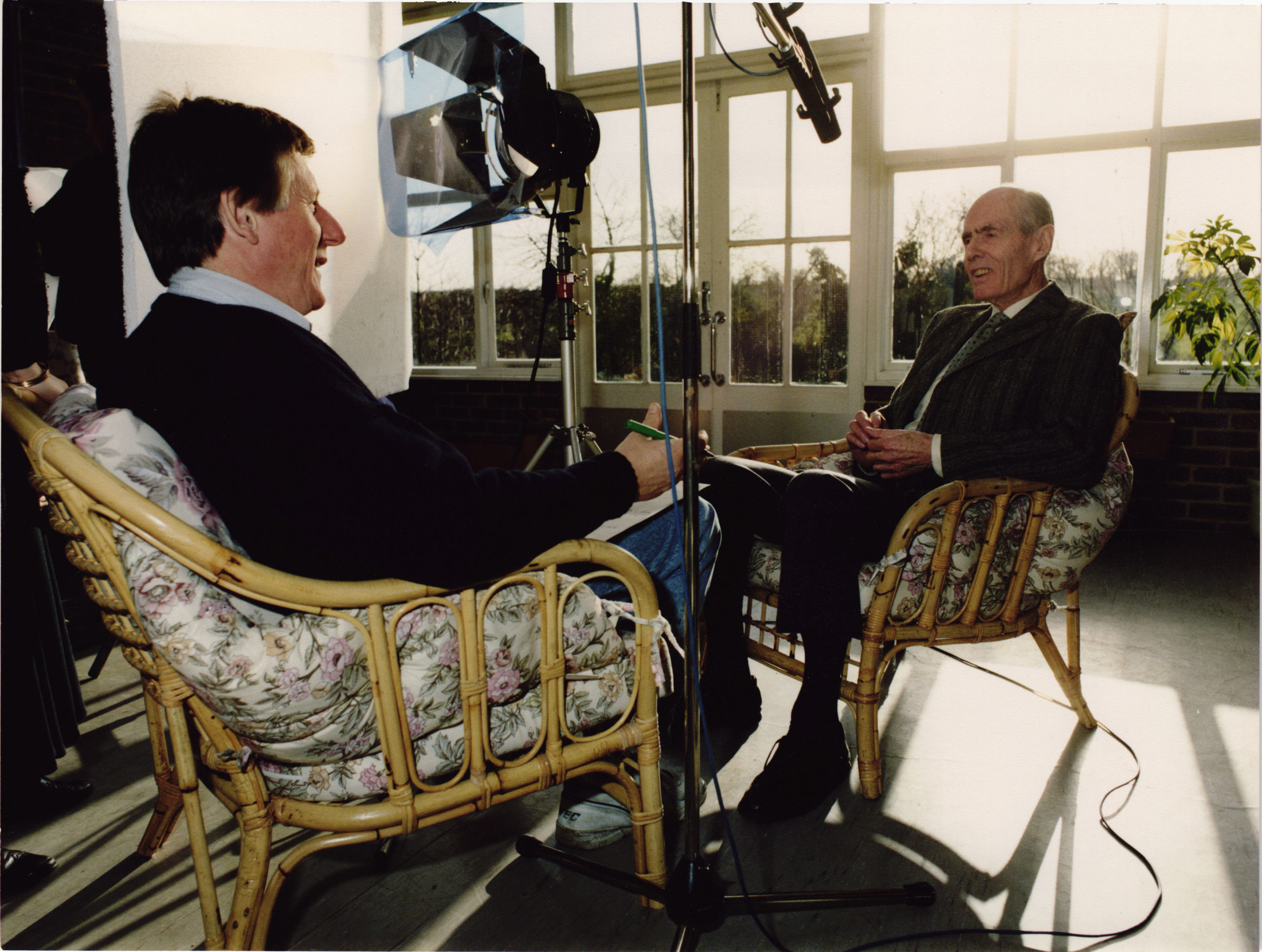 A man being interviewed, sat in a conservatory surrounded by microphones and lights